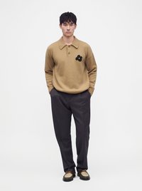 Jeune homme debout, mains dans les poches, portant un pull beige avec un détail de fleur noire, un pantalon noir et des mocassins bicolores sur fond blanc.