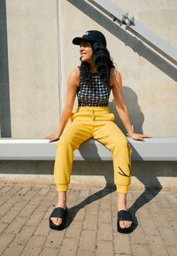 Pantalon de survêtement jaune avec des accents noirs, crop top à motifs noirs et blancs, et sandales noires à enfiler. Assis sur un banc en béton.