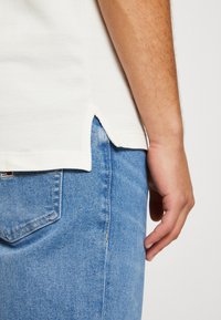 Camiseta blanca con aberturas laterales, combinada con jeans de mezclilla azules. La textura de la camiseta es suave, y los jeans tienen un lavado claro.