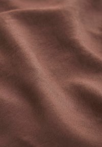 Tessuto marrone con una superficie liscia, caratterizzato da leggere pieghe che creano profondità e variazioni di colore sulla superficie del materiale.
