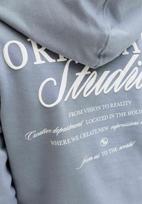 Tuva zila kapučveida džempera auduma ar baltu izdrukātu tekstu dažādos burtu šrifta stilā par radošu studiju un vīziju īstenošanā.