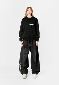 Sudadera negra con logo blanco en el frente, combinada con pantalones de mezclilla oversize negros que presentan un gráfico lateral. Zapatos chunky de color claro.