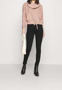 Rosa gestrickter Pullover mit Rollkragen, der an der Taille gebunden ist. Schwarze enge Jeans und schwarze Stiefel mit Absatz. Hält eine cremefarbene Handtasche.