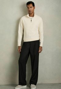 Cremfarbener Reißverschluss-Sweatshirt und schwarze locker sitzende Hose, dazu hellgraue Sneakers. Glatte Stofftextur mit entspanntem Schnitt.