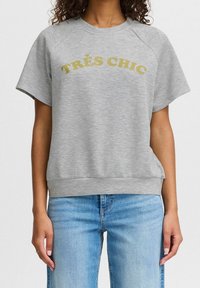 Grå sweatshirt med korta raglanärmar, ribbad nederkant och "TRÈS CHIC" tryckt i guld. Tillverkad av mjukt, texturerat tyg. Kombinerad med ljusblå jeans.
