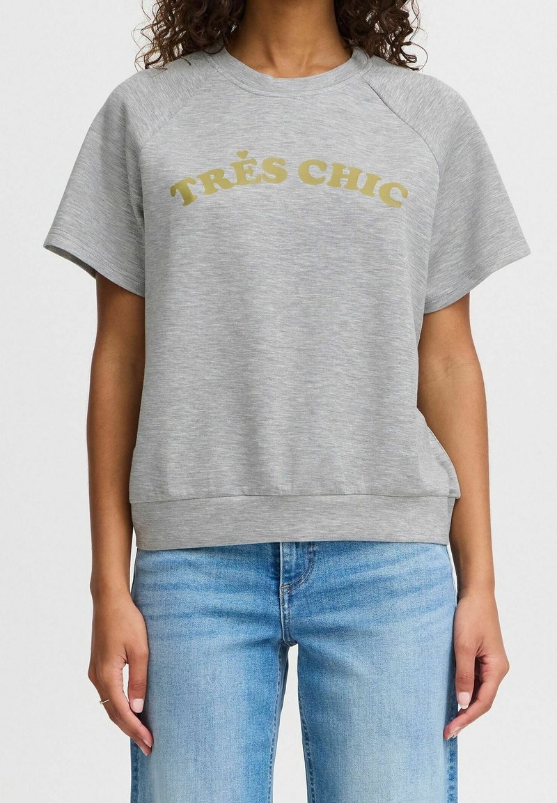 Grå sweatshirt med korta raglanärmar, ribbad nederkant och "TRÈS CHIC" tryckt i guld. Tillverkad av mjukt, texturerat tyg. Kombinerad med ljusblå jeans.