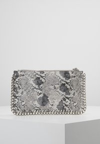 Hedvábný obdélníkový psaníčko s šedým a černým hadím vzorem, se stříbrným řetízkem, vystavené na bílém podstavci na jednobarevném pozadí.