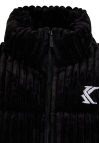 Black plush vest featuring ribbed texture, high collar, and front zipper. Embroidered logo in white on the left side.