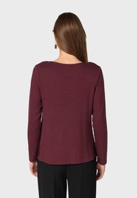 Burgundowa bluzka z długim rękawem, okrągłym dekoltem, rozcięciami po bokach u dołu i miękkim, teksturowanym materiałem. Noszona z czarnymi spodniami.