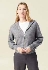 Grå zip-up hoodie tillverkad av texturerat tyg, med en huva, ribbade muddar och en subtil glans på dragkedjan. Matchad med en vit skjorta.