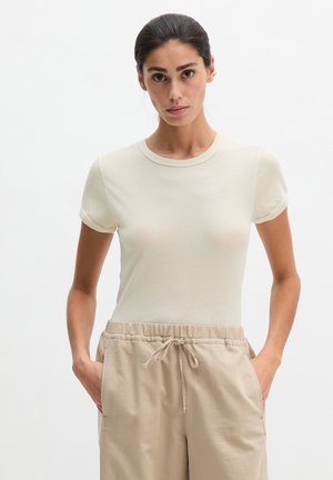 Vrouw met donker haar, gekleed in een beige top met korte mouwen en lichtbruine broek met tunnelkoord, staande met handen in de zakken tegen een witte achtergrond.