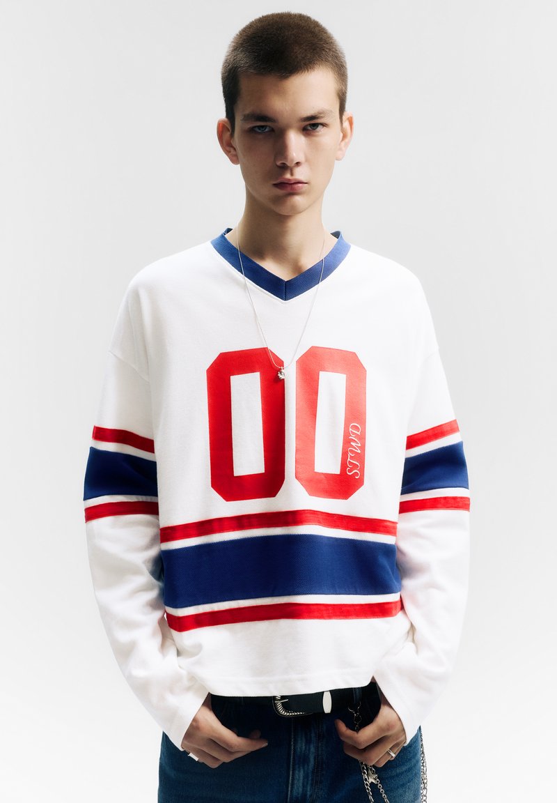 Biała koszula z długim rękawem z czerwonym nadrukiem "00", niebiesko-czerwonymi paskami na rękawach i dekoltem w serek. Noszona z niebieskimi dżinsami i łańcuchem.