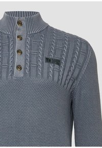 Szary sweter wykonany na drutach z wysokim kołnierzem, czterema ciemnymi guzikami, wzorem w warkocz oraz małą naszywką z logo "Elevated Casual Est. 1969".