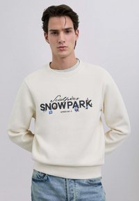 Sudadera blanca con cuello redondo con gráfico de "SNO PARK", que presenta ilustraciones coloridas con temática de esquí. Combina con jeans azules. Textura suave.