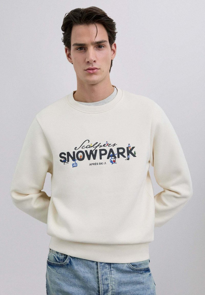 Sudadera blanca con cuello redondo con gráfico de "SNO PARK", que presenta ilustraciones coloridas con temática de esquí. Combina con jeans azules. Textura suave.