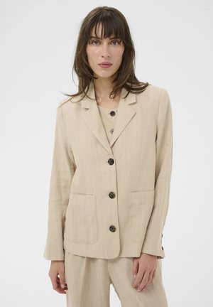 Femme portant un blazer beige à fines rayures avec quatre boutons, deux poches avant, et un pantalon large assorti sur un fond blanc uni.