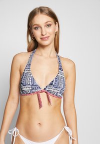 Mørkeblå halterneck bikini topp med hvite og røde tribal mønstre; har en dyp V-design og en frontknute lukking. Glatt stoffoverflate.