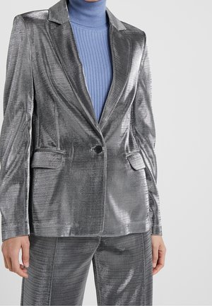 Frau trägt einen glänzenden silbernen Blazer und passende Hosen über einem gerippten blauen Rollkragenpullover, stehend vor hellem Hintergrund.