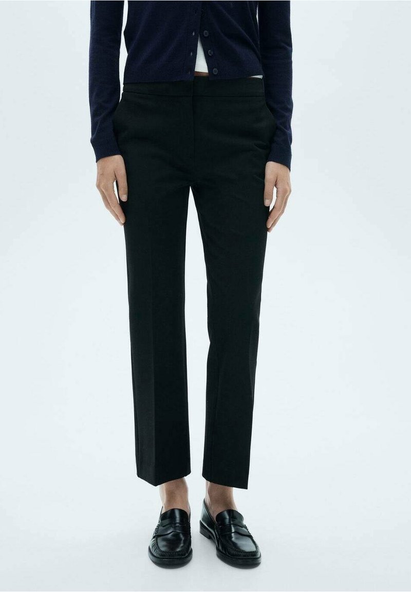 Personne portant un pantalon noir droit, des mocassins noirs sans lacets et un cardigan sombre boutonné, debout devant un fond clair uni.