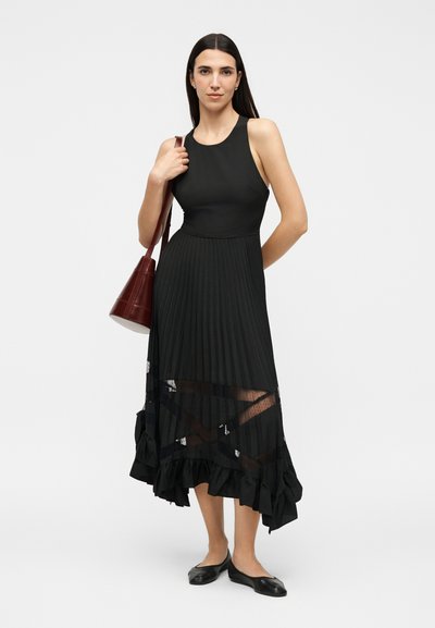 Γυναίκα με μακριά σκούρα μαλλιά που φοράει αμάνικο μαύρο πλισέ μίντι φόρεμα με διαφανείς λεπτομέρειες και μαύρα φλατ παπούτσια, κρατώντας μια σκούρα καφέ δερμάτινη τσάντα ώμου.