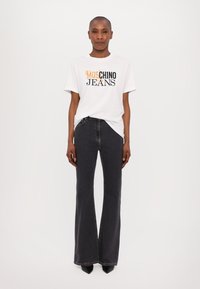 Bílý tričko s černým a oranžovým logem "MOSCHINO JEANS", v kombinaci s černými rozšířenými džíny. Modelka má na sobě špičaté černé podpatky.