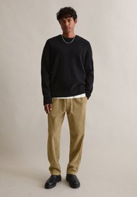 Μαύρο πλεκτό πουλόβερ, κρεμ πουκάμισο από κάτω, χακί παντελόνι από κοτλέ και μαύρες μπότες αστραγάλου. Χαλαρή εφαρμογή με υφασμάτινη υφή.