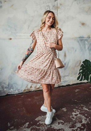 Květinové šaty v světle růžové barvě s volánovými rukávy a elastickým pasem. Spárovány s bílými teniskami. Jednoduchá crossbody taška. Texturované pozadí.