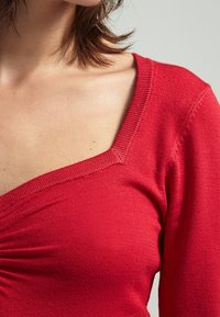 Top rosso aderente con un ampio scollo quadrato, maniche lunghe e dettagli arricciati sul davanti. Realizzato in morbido e liscio tessuto a maglia.