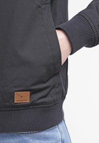 Svart bomberjacka med ribbade ärmslut och nederkant, i ett strukturerat tyg. Brunt läderlogotypmärke på nedersta vänstra sidan nära fickan.