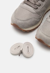 Paire de baskets en daim gris avec tissu en mesh et lacets beiges clairs enroulés sur une surface blanche.