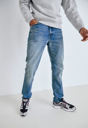 Jeans de mezclilla azul con un acabado gastado, corte recto y diseño de cinco bolsillos, combinados con zapatillas deportivas en blanco y negro. Sudadera gris visible.