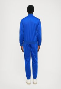 Man standing facing away wearing a blue tracksuit with black stripes on sleeves, white shoes, on a plain light background.