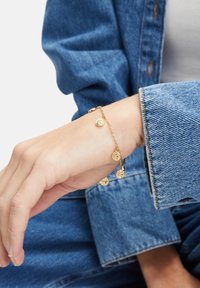 Goldener Charm-Armband mit runden Anhängern mit strukturierten Designs, gesichert mit einem Verschluss, getragen an einem Handgelenk mit Jeanskleidung.