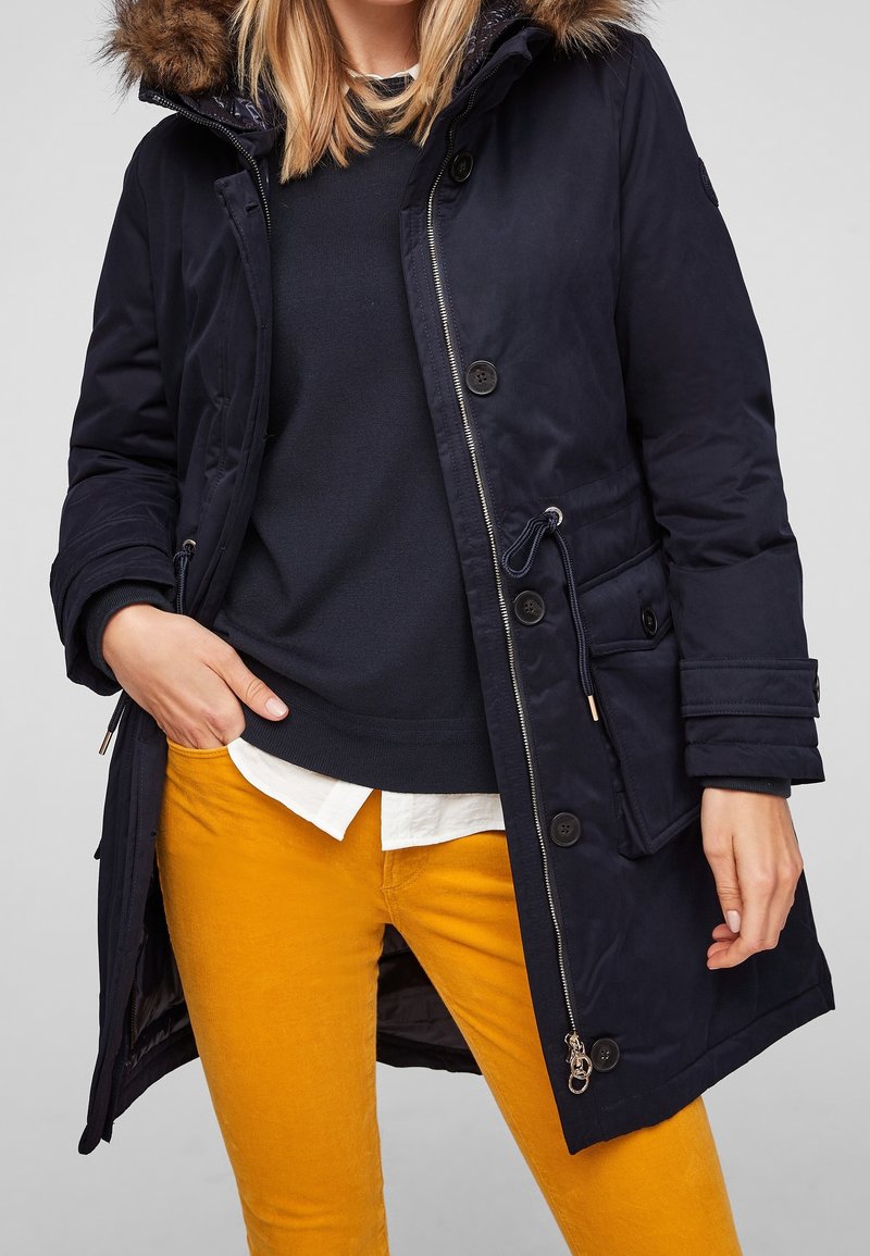 Parka bleu marine avec capuche en fausse fourrure, fermeture éclair et boutons, taille ajustable et poches latérales. Associée à un pantalon en velours côtelé jaune.