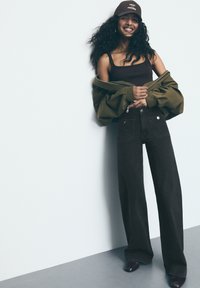 Schwarze weit geschnittene Jeans mit Fronttaschen, kombiniert mit einem braunen Tanktop und einer olivgrünen oversized Jacke sowie einer braunen Mütze mit weißem Text.