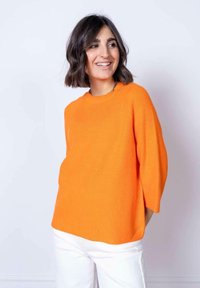 Maglione a costine arancione con scollatura rotonda e maniche a tre quarti, abbinato a pantaloni bianchi. Tessuto liscio, vestibilità casual.