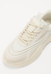 Hvid tekstureret sneaker med lagdelt design, der har øverdel af mesh, tykkeste cremefarvede sål og tonede snørebånd. Bemærkelsesværdige riller langs siden.