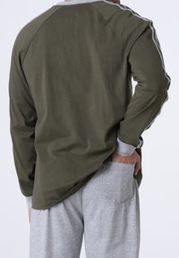 Camicia a maniche lunghe verde oliva con colletto grigio e righe a contrasto sulle maniche, abbinata a pantaloni jogger grigi chiari con tasche.