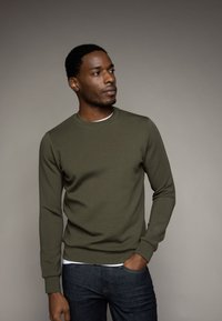 Sudadera verde oliva de manga larga y escote redondeado, hecha de una tela suave. Combinada con jeans de color azul oscuro.