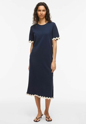 Femme debout portant une robe midi bleu marine avec une bordure ondulée crème sur les manches et l'ourlet, associée à des sandales à talons noires ouvertes à l'avant.