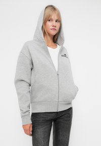 Siva zip-up kapuljača s kapuljačom i logotipom na prsima. Ima rebraste manžete i rub, izrađena je od mekanog materijala, u kombinaciji s tamnim hlačama.