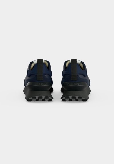 Paire de baskets bleu marine avec semelles épaisses noires vues de dos sur un fond gris clair uni.