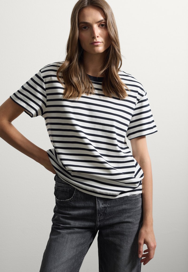 Gestreepte t-shirt met korte mouwen in wit en marineblauw. Katoenen stof, relaxte pasvorm, ronde hals. Gecombineerd met donker gewassen jeans.
