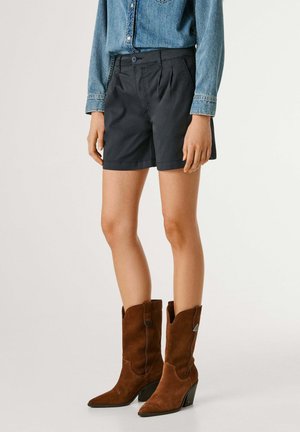 Person trägt schwarze Shorts, ein langärmeliges Denim-Hemd und braune Wildleder-Cowboystiefel bis zur Mitte der Wade und steht vor einem einfachen Hintergrund.