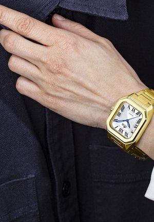Orologio quadrato tone oro con quadrante bianco, lancette blu e numeri romani. Indossato su un polso con una camicia blu con bottoni.