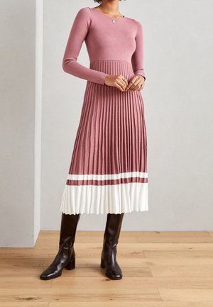 Kvinde iført en lyserød kjole med lange ærmer og et plisseret skørt med hvide og lyserøde striber, kombineret med sorte støvler, der går op til midt på læggen, stående på et trægulv.