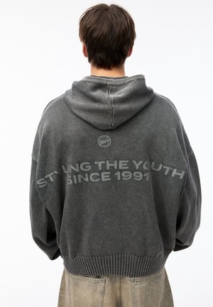 Osoba z krótkimi brązowymi włosami nosi oversize szarą bluzę z kapturem z napisem „STYLING THE YOUTH SINCE 1991” na plecach, w połączeniu z beżowymi spodniami.