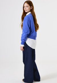 Sudadera azul sobre una camisa blanca con cuello, combinada con pantalones de mezclilla oscura de pierna ancha. El cabello lacio y largo cae más allá de los hombros.