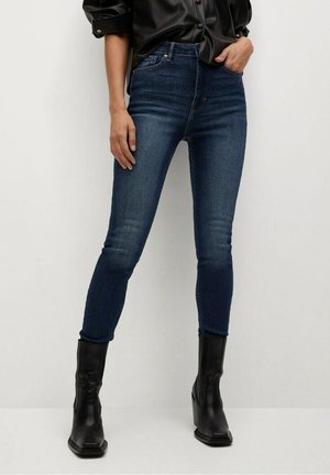 Kvinde iført mørkeblå skinny jeans og sorte læderankelstøvler, stående med den ene hånd i lommen mod hvid baggrund.