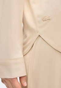 Μπεζ μακρυμάνικο πάνω μέρος με μανσέτα, λεπτομέρεια πλαϊνής σχισμής και μικρό κεντημένο λογότυπο μάρκας κοντά στη μέση.